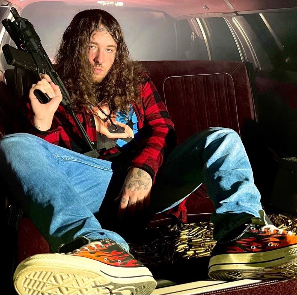 Reaper Man sitting in the back of a car in a red flannel shirt, jeans and flame sneakers